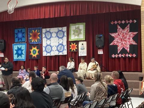 Students sitting on the stage in front of quilts showing off their Cultural clothing