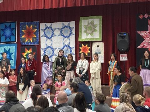 Students standing on the stage in front of quilts showing off their Cultural clothing