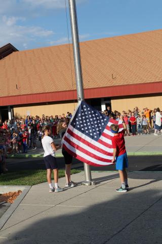 Two Fifth-grade students holding the United States Flag while one student runs it up the flag pole