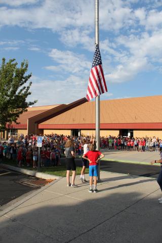 One student running the United States Flag up the pole, in the back ground students from the whole school stand at attention
