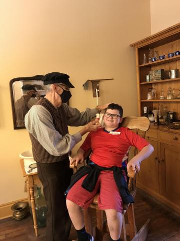 A student sitting in a barber's chair as a gentleman shows what it would be like to get a shave