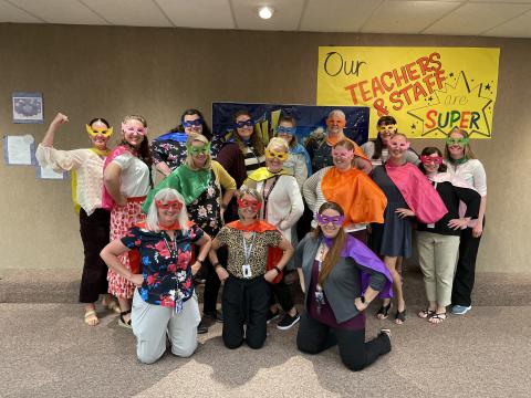 Teachers and Staff with Super Hero capes and masks on in front of a sign that says "Our Teachers and Staff are Super"