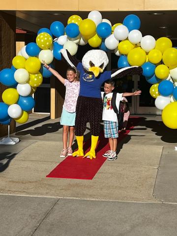 The Art City Eagle standing with students on the red carpet under the balloon arch