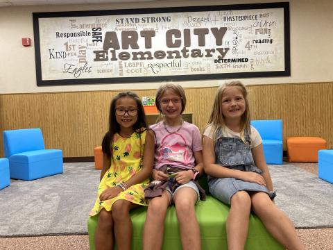 Three girls sitting on a bench in front of the Art City Elementary picture