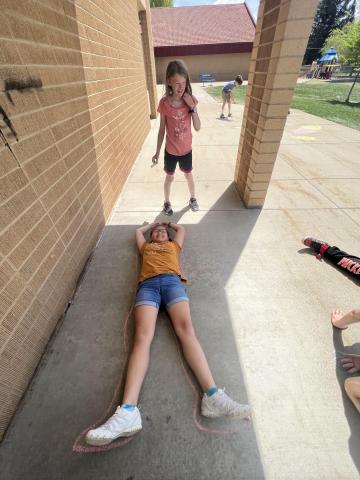 one student laying on the ground with arms outstretched, while another student traces around them. 