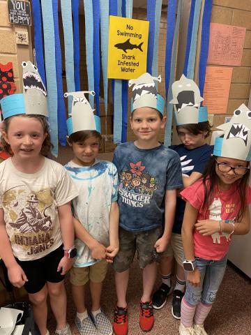Five students standing in front of "No Swimming, Shark Infested Water" with Shark heads on their head