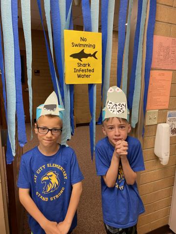 Two students standing in front of "No Swimming, Shark Infested Water" with Shark heads on their head