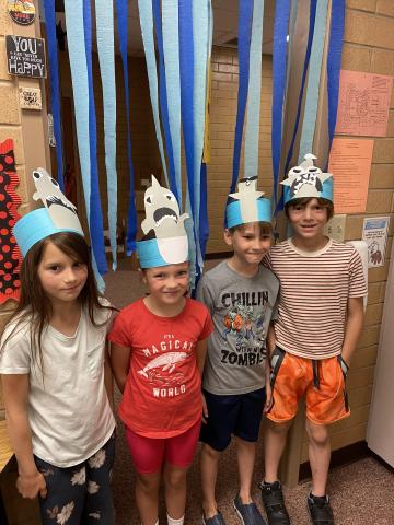 Four students standing in front of "No Swimming, Shark Infested Water" with Shark heads on their head