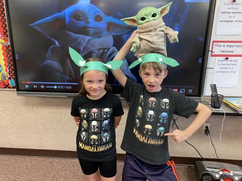 Two students with Yoda ears in front of a picture of Grogu and one student holding a stuffed Grogu on his head.