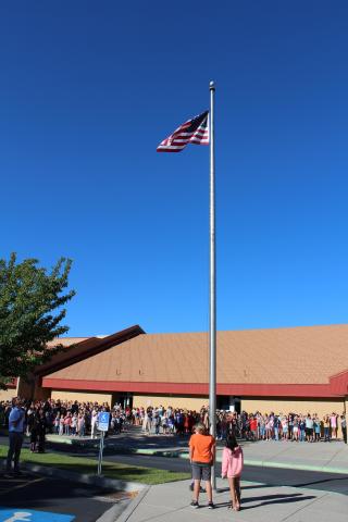 Three fifth-grade students lowering the flag to half staff with the wind blowing the flag out and Art City Student body in the background