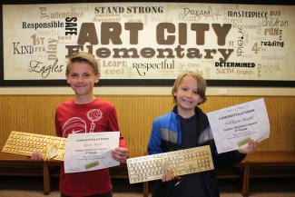 One fourth grade student and one fifth grade student holding a certificate of award for top finishers and golden keyboards