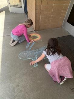 Two students drawing and coloring, with chalk, a rabbit and a sunshine on the sidewalk. 